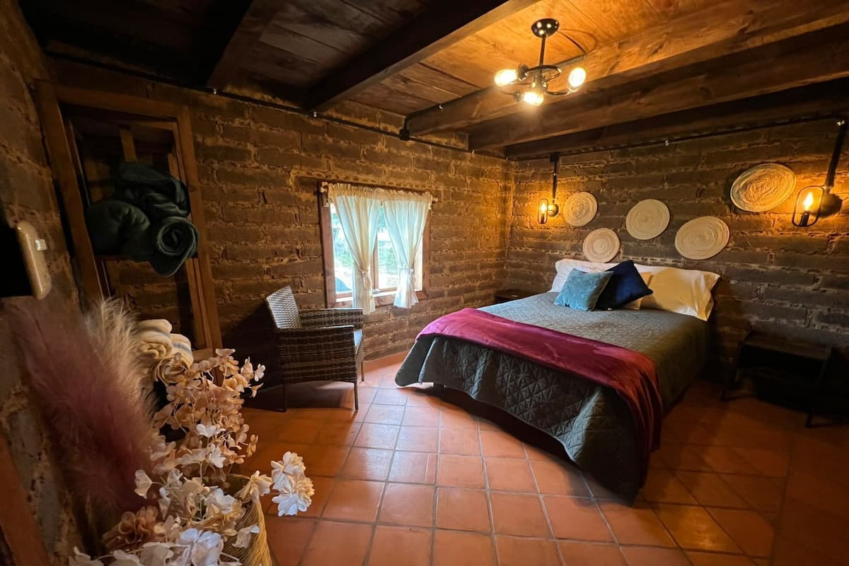 A rustic wooden room with a bed, a wicker chair, and a window with curtains.