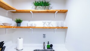 Une cuisine avec des plans de travail blancs, un évier en acier inoxydable et des étagères en bois où sont disposées des plantes et des ustensiles de cuisine.