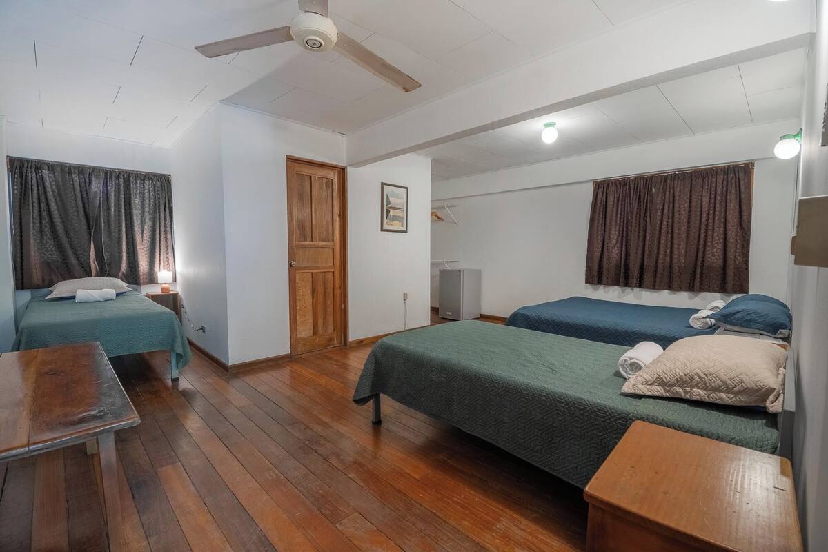 A room with two single beds, a wooden floor, a ceiling fan, a small table, and a door.