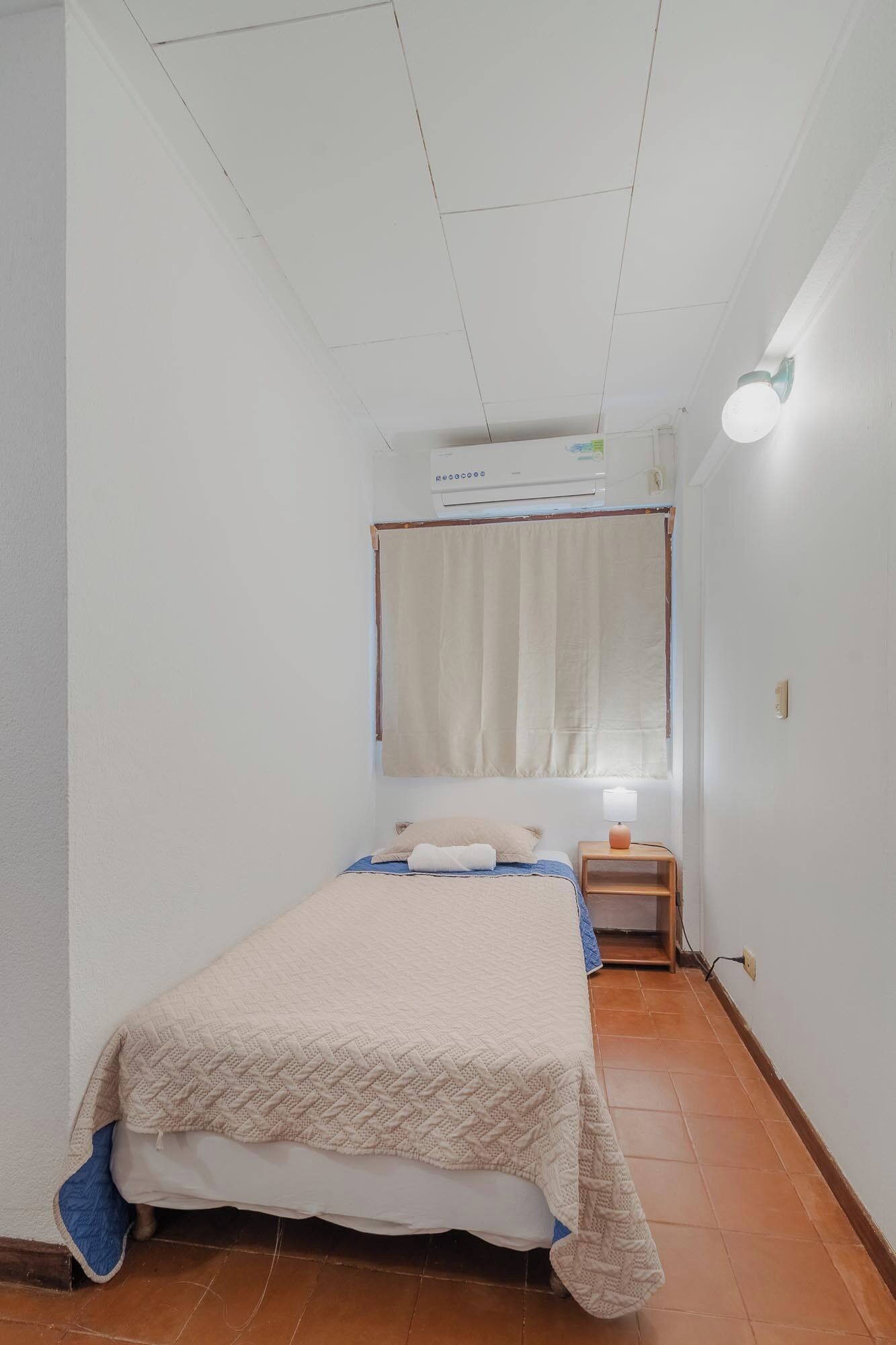 A small, narrow room with a single bed, a nightstand, a wall-mounted air conditioner, and a ceiling light.