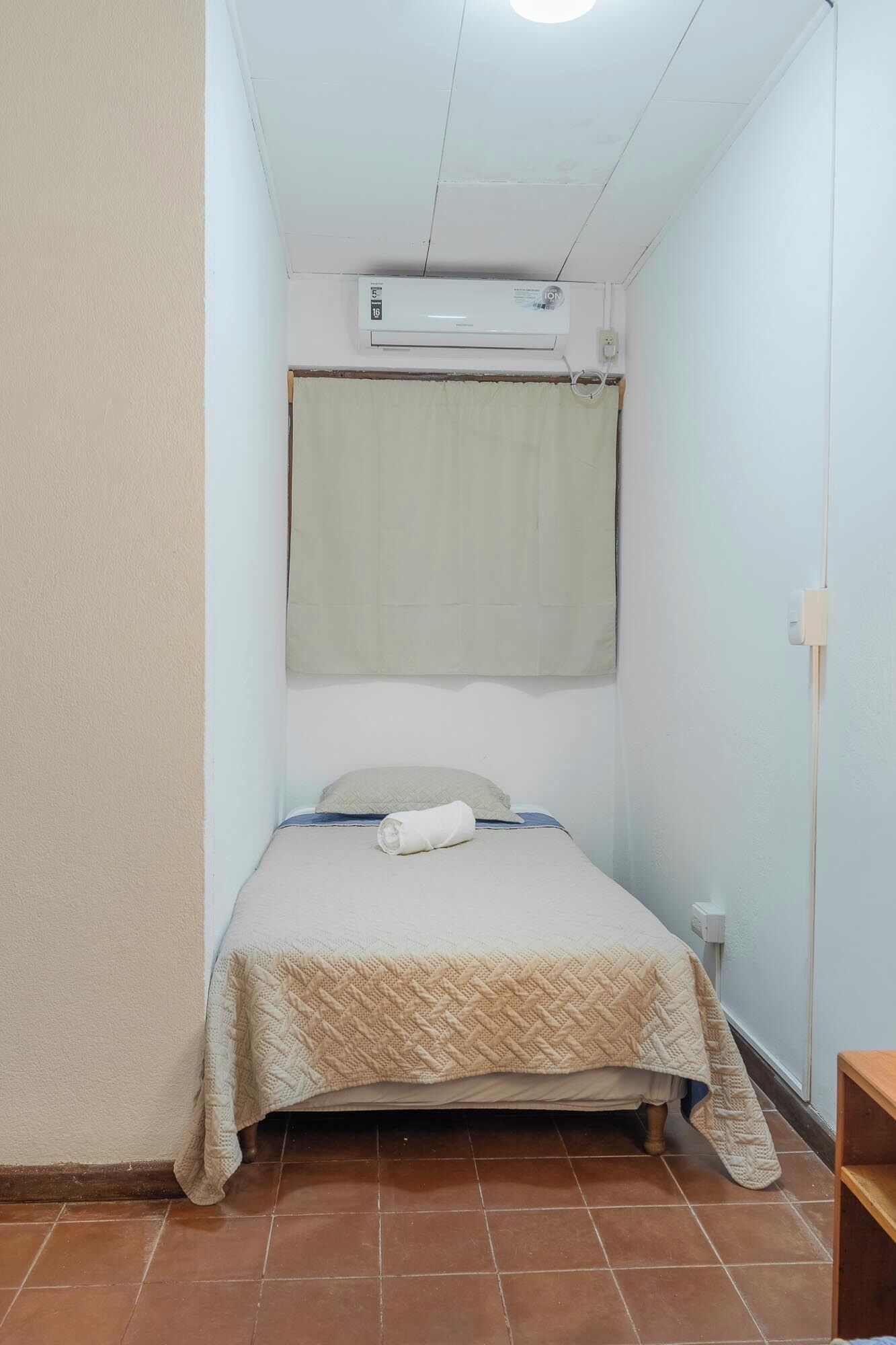 A small, single-bed room with a white air conditioning unit above the bed, a plain wall, and a tiled floor.