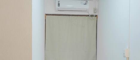 A small, single-bed room with a white air conditioning unit above the bed, a plain wall, and a tiled floor.