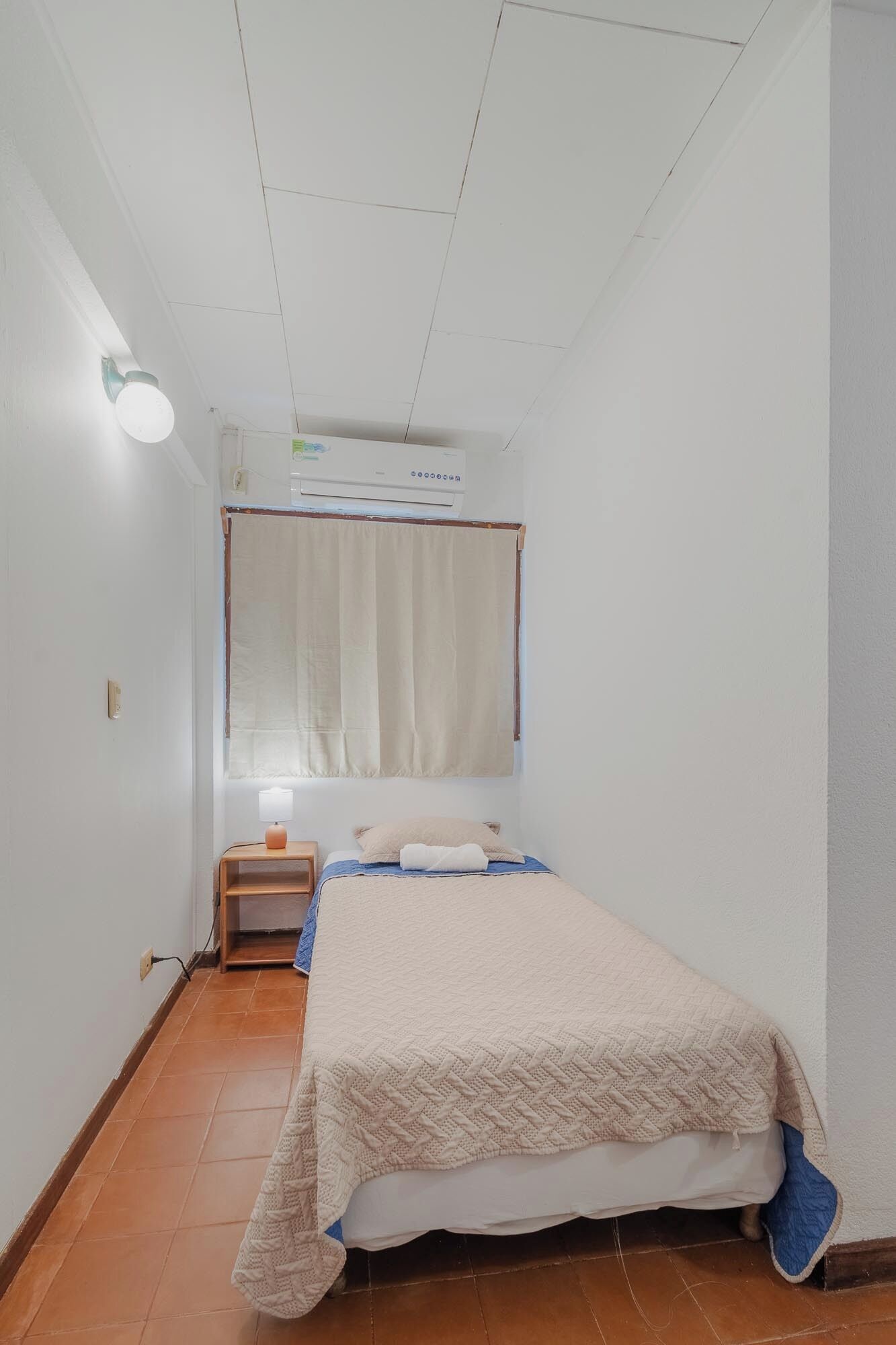 Quarto pequeno e limpo com cama, mesa de cabeceira, luminária e janela com cortinas.
