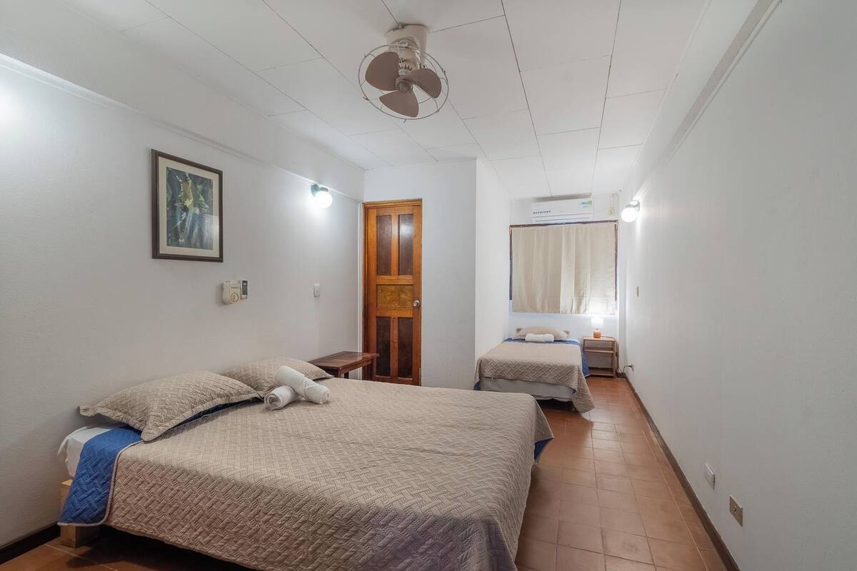 A hotel room with two beds, a ceiling fan, a wooden door, a small table, and a bedside table with a lamp.