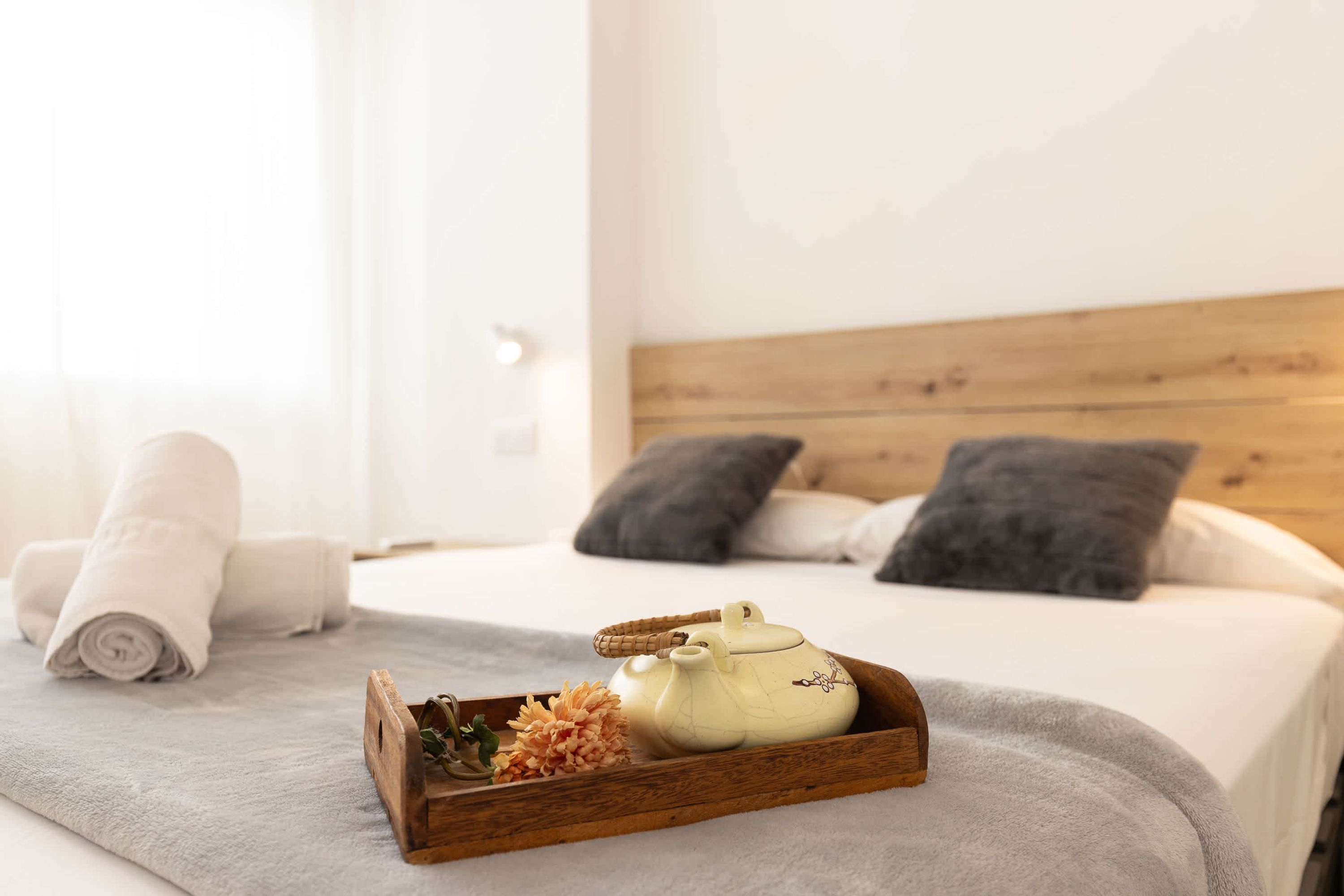 Ein ordentlich bezogenes Bett, gerollte weiße Handtücher, ein Holztablett mit Teekanne und Blumen sowie eine graue Decke.