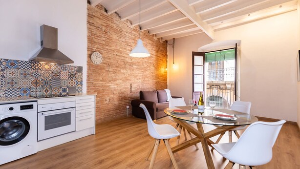 A modern kitchen with a washing machine, a dining area with a glass table and white chairs, a brick wall, and a hanging light fixture. - Merceria 1 by Unik Tarragona (Tarragona)