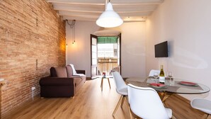A modern living room with a brick wall, wooden flooring, a glass dining table, white chairs, a sofa, and a TV mounted on the wall. - Merceria 1 by Unik Tarragona (Tarragona)