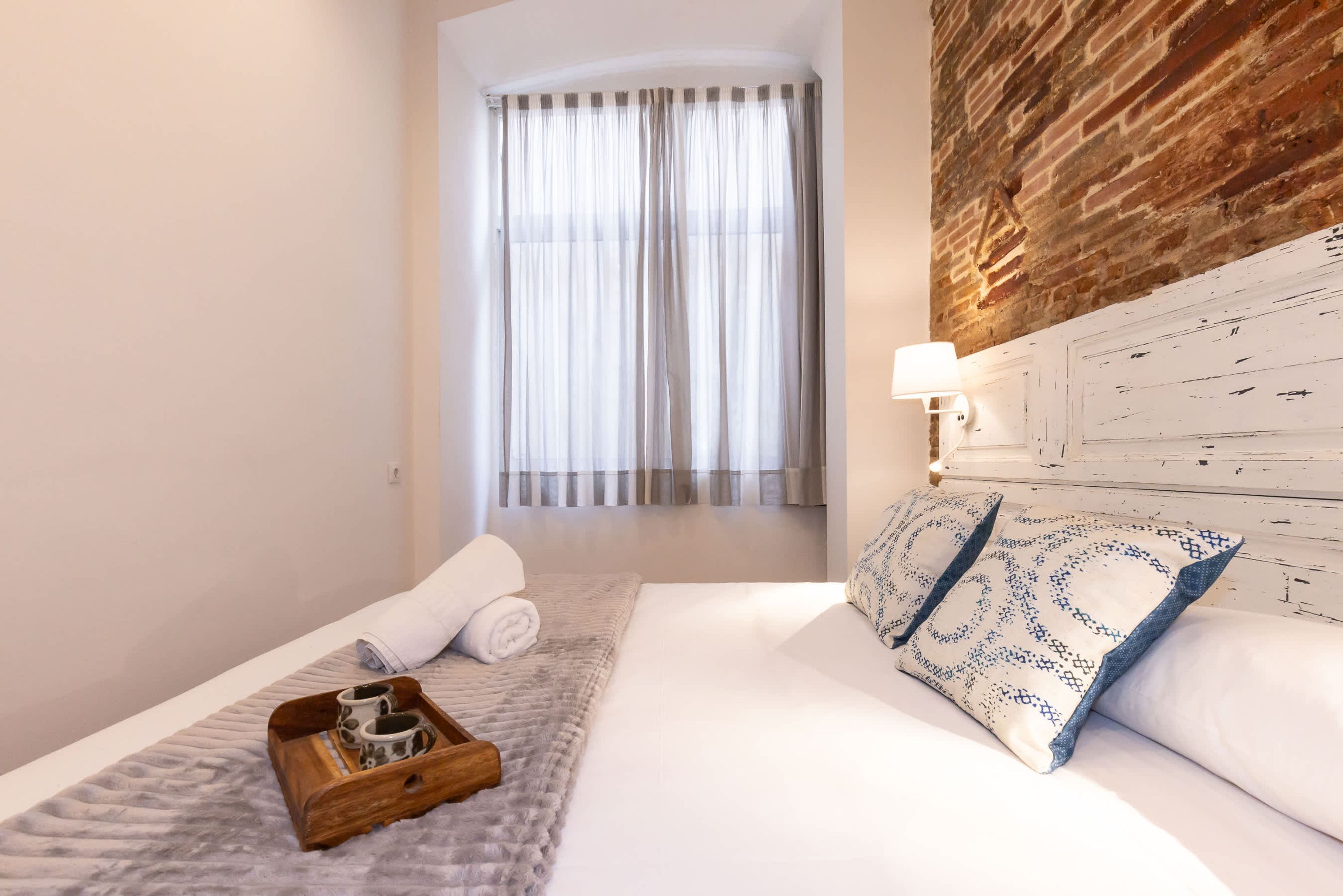 A neatly made bed with a wooden tray, a rolled towel, and a patterned pillow. A window with sheer curtains is visible in the background.