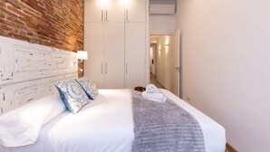A bedroom with a brick accent wall, a white headboard, a bed with white linens, a grey throw blanket, and a white towel. - Merceria 1 by Unik Tarragona (Tarragona)