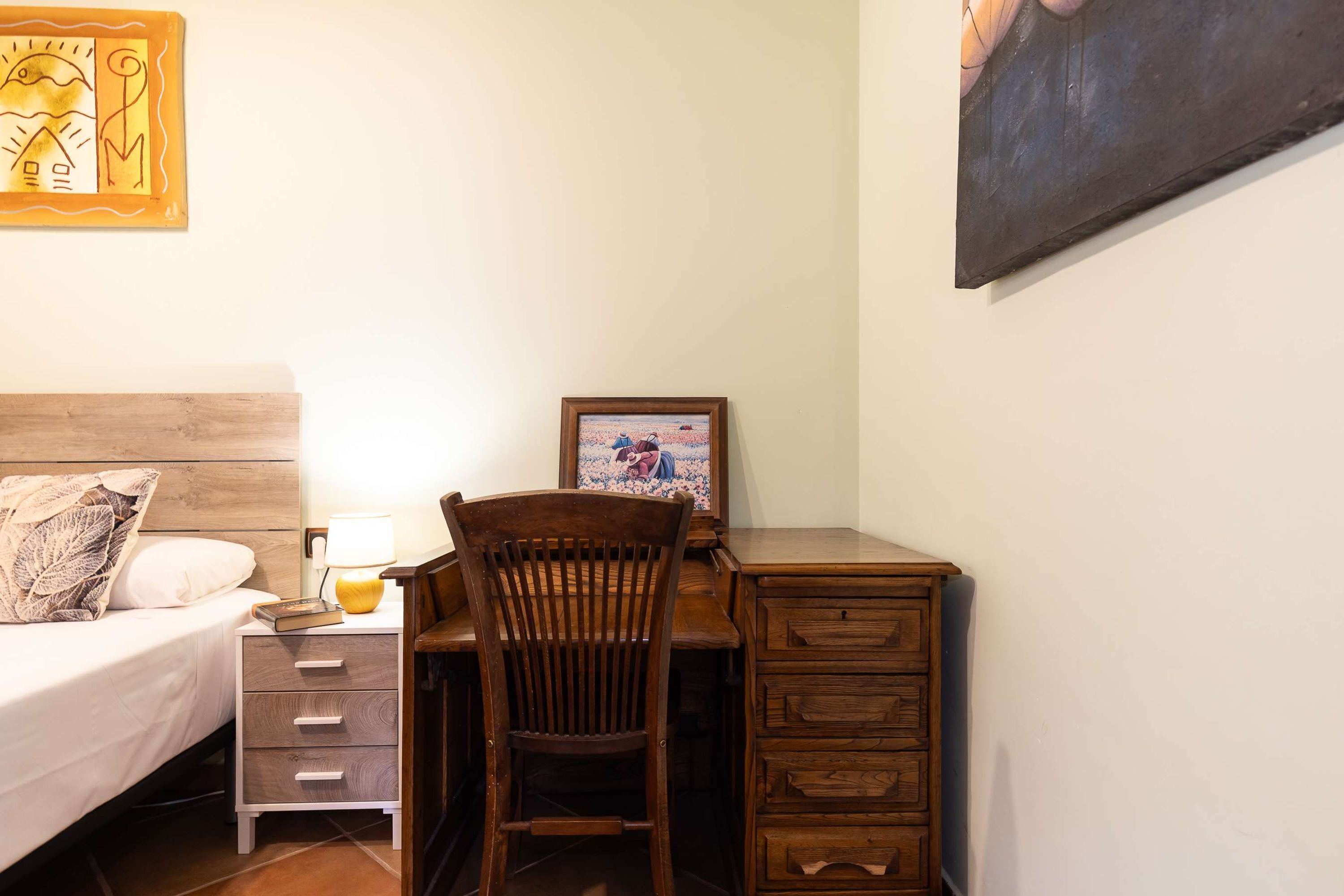 Un dormitorio con cama, mesita de noche, escritorio, silla y un cuadro en la pared.