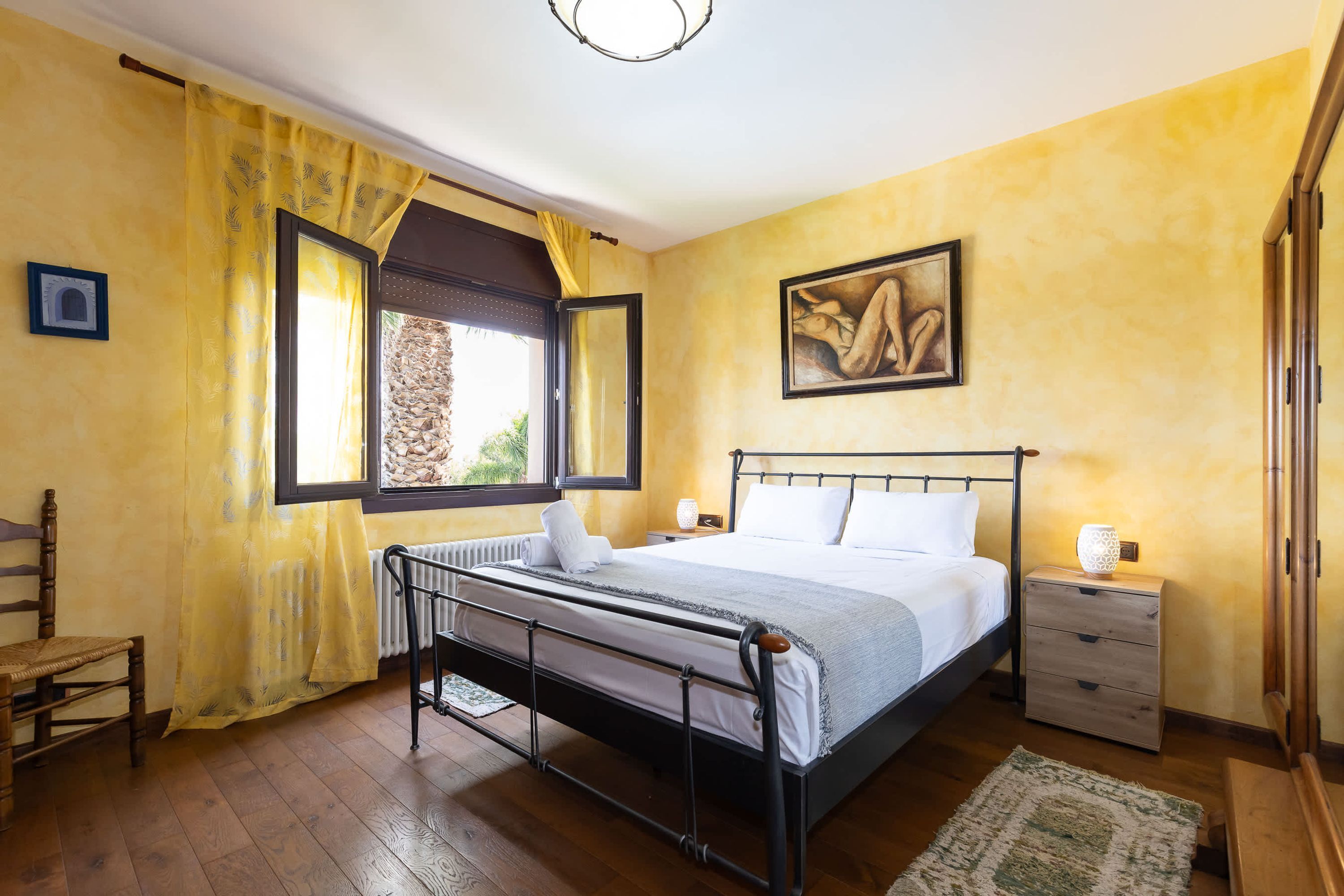 Une chambre à coucher avec un lit, une table de chevet, une chaise, une fenêtre avec des rideaux et un tableau au mur.