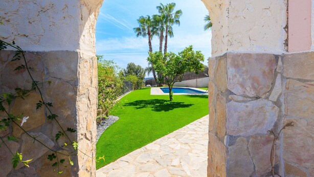 Un arcoado acceso de piedra que da a un césped bien cuidado con una palmera y una piscina.