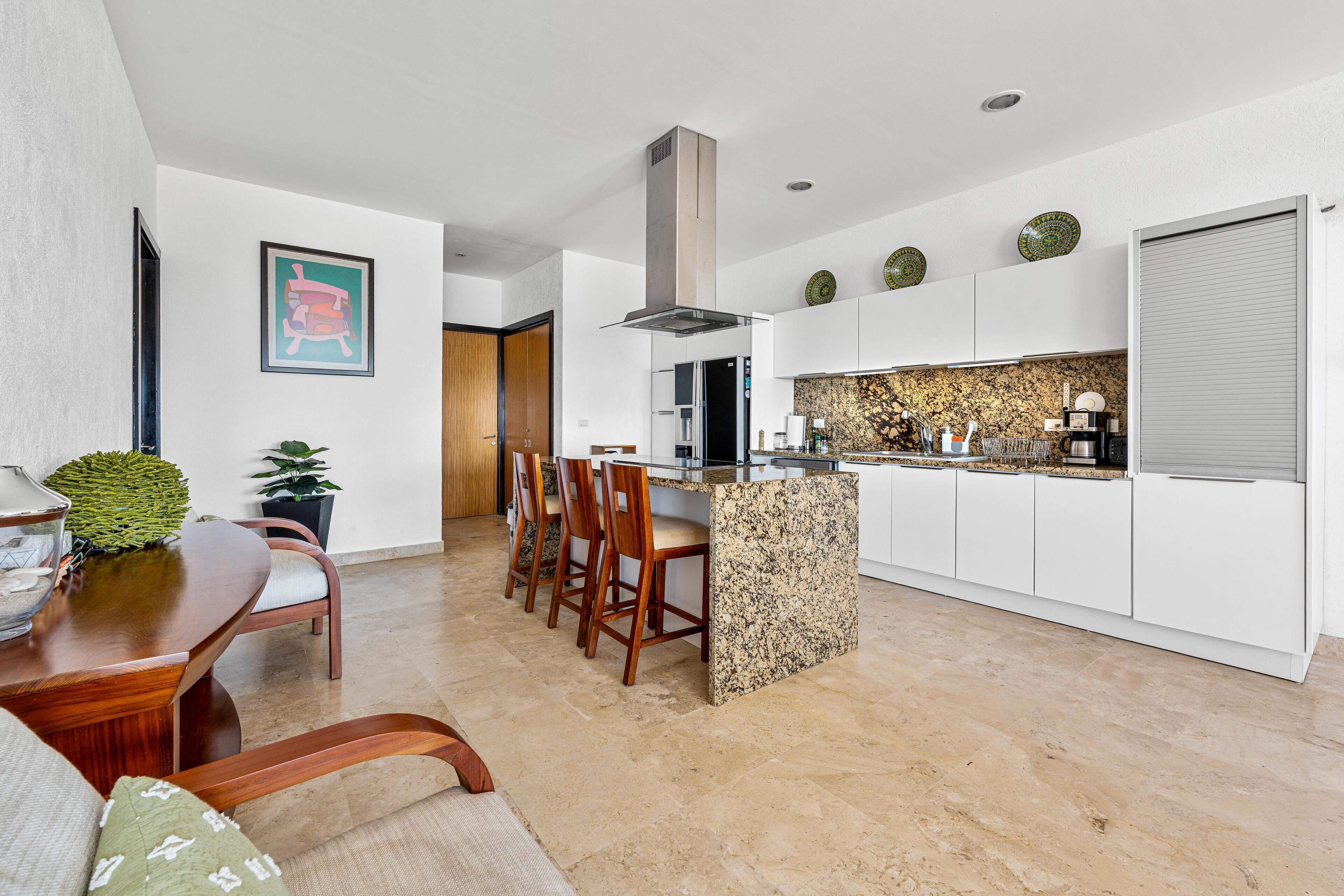 A modern kitchen with white cabinets, a granite island, and a dining area with wooden chairs and a table.