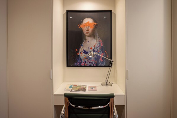 Un escritorio con una obra de arte enmarcada, una lámpara y una silla en una habitación con paredes lisas.