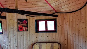 Een houten blokhut van binnen, met een bank, twee stoelen, een kleine tafel en een raam met rode randen.