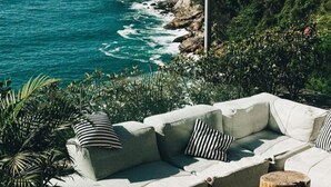 A coastal outdoor seating area with a white sofa and striped cushions, surrounded by greenery and overlooking the ocean. -  Luxury Ocean Front Loft in Rio De Janeiro Joa (Joá Rio de Janeiro)