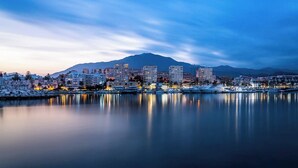 A coastal cityscape at dusk with buildings reflecting on calm waters, a marina with boats, and a mountain in the background.
