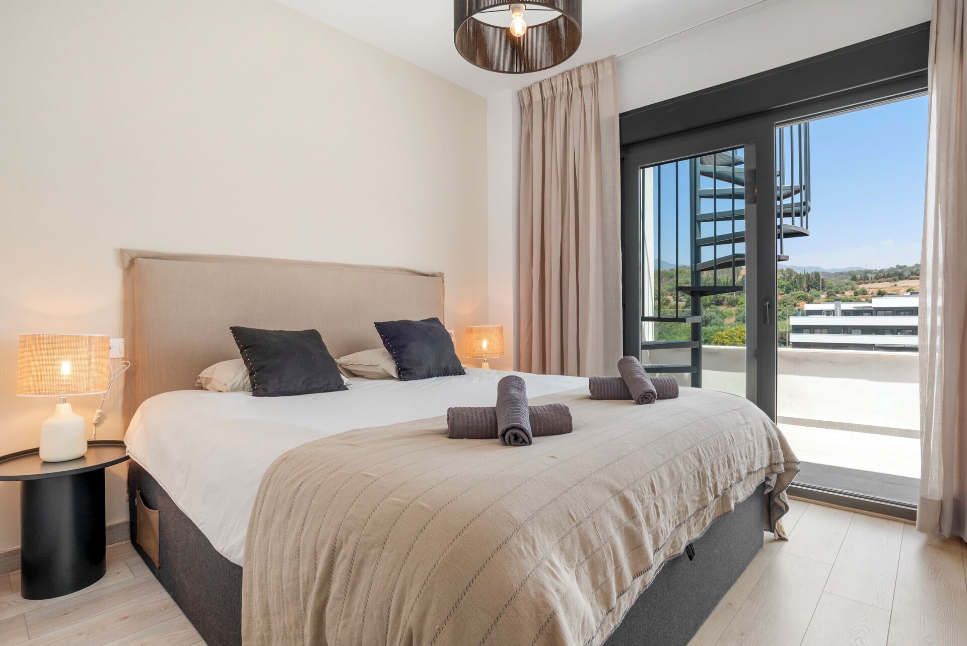 A modern bedroom with a large bed, two bedside tables with lamps, and a view of a balcony with a spiral staircase.