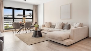 A modern living room with a beige sofa, a round coffee table, and a dining area with chairs and a table. Large windows offer a view of the cityscape.