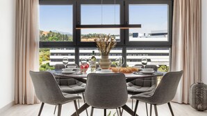 A dining area with a table set for four, large windows with a view of buildings and greenery, and a vase with dried flowers on the table.