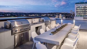 Eine Dachterrasse mit Grill, Barhocker und Blick auf die Dämmerung über der Stadt.