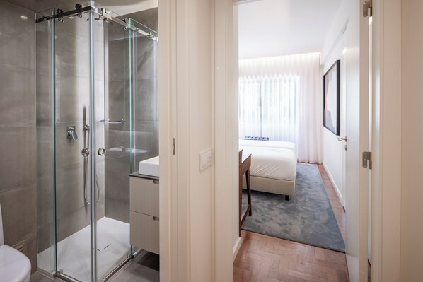 A modern hotel room with a glass-enclosed shower, a bed with white bedding, and a large window with sheer curtains.