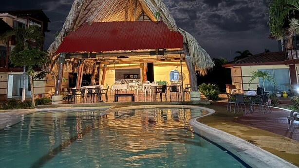 Un edificio con techo de paja, con piscina delantera, rodeado de palmeras y otras construcciones.