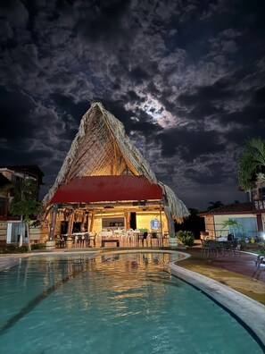 A thatched-roof building with a pool in front, surrounded by palm trees and other buildings. - Hotel Bella Suiza Campestre  (Villavicencio)