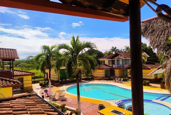 A swimming pool with lounge chairs and a palm tree, surrounded by buildings with a thatched roof. - Hotel Bella Suiza Campestre  (Villavicencio)