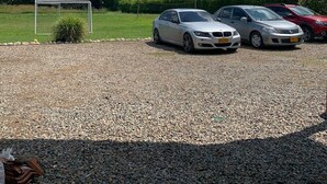 Un estacionamiento con suelo de grava, un arco de fútbol y tres coches aparcados bajo una zona techada con techo de paneles ondulados.