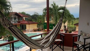 Una hamaca en un balcón con baldosas, con piscina y sillas, con vistas a una zona residencial.
