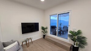 Una sala de estar con televisor de pantalla plana, un sofá gris, una mesa de centro de vidrio y un ventanal corredero que da a un patio exterior con sillas y una mesa.