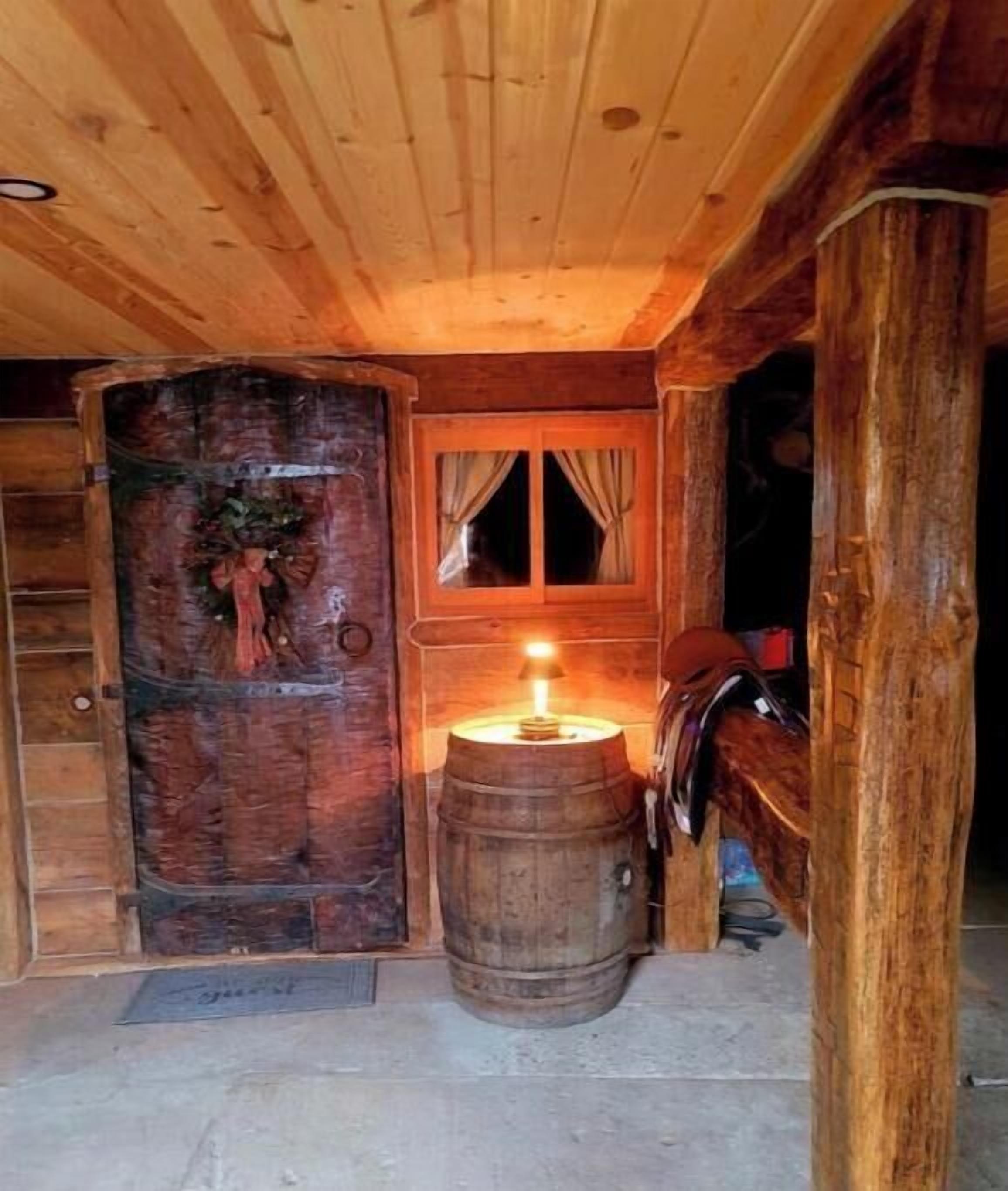 Interno di una rustica baita di legno, con una porta in legno, una finestra con tende e una candela accesa su una botte.