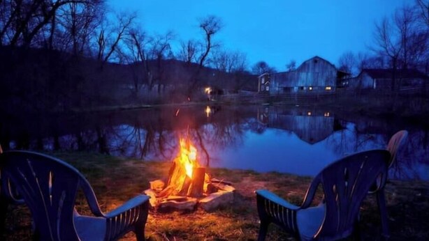A campfire with two chairs facing it, a calm lake, and a house in the background. - Rustic Luxury w/Horses-Historic Whiskey Distillery (Watsontown)