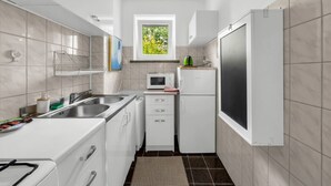 Cocina compacta con muebles blancos, microondas integrado, refrigerador y ventana con vistas a vegetación.