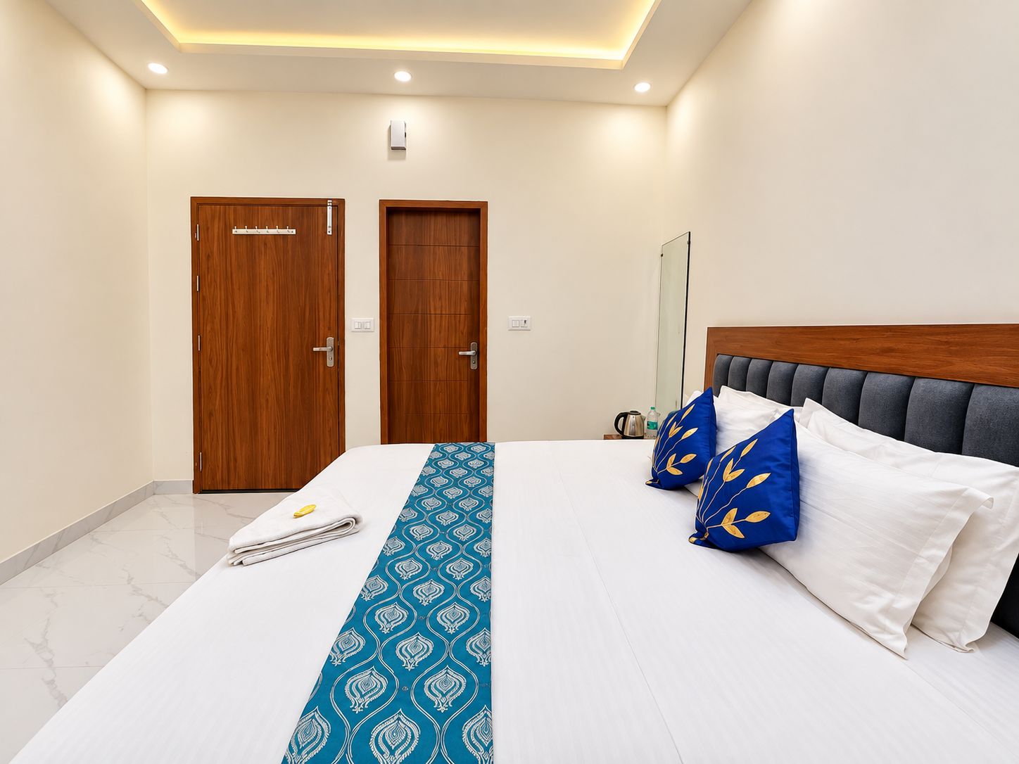 A hotel room with a bed, a wooden wardrobe, a mirror, and a bedside table with a kettle and a bottle.