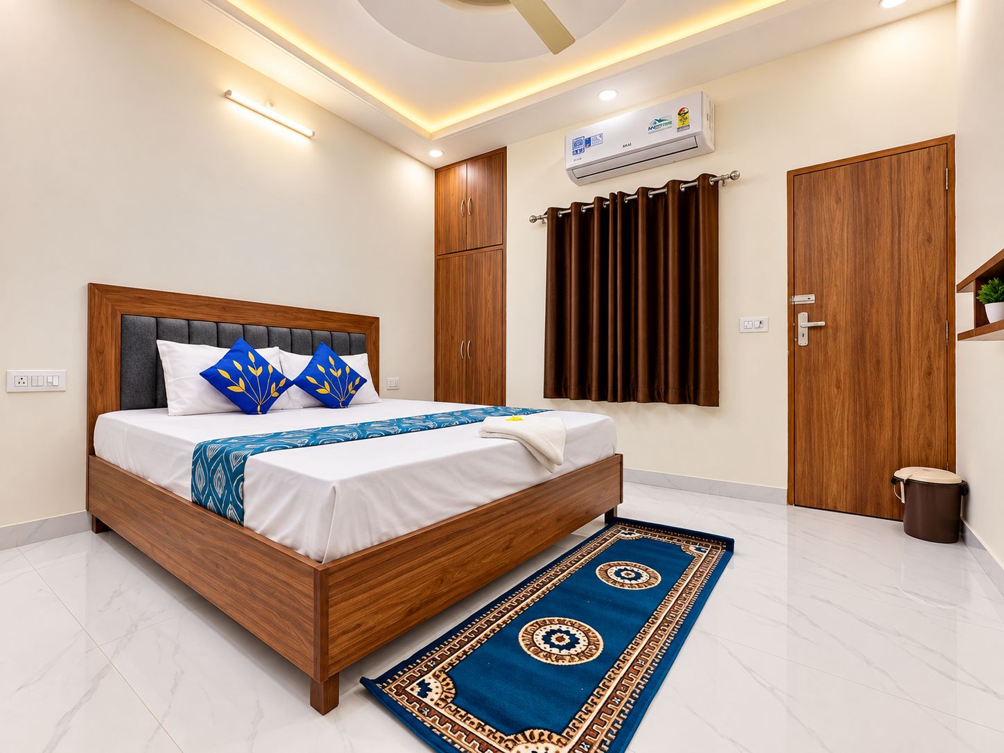 A modern bedroom with a wooden bed, a blue patterned bedspread, a white wall, a ceiling light, a wall-mounted air conditioner, and a brown door.