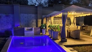 A swimming pool with blue lighting, a covered outdoor seating area with tables and chairs, and potted plants. - Bryanston Palms Accommodation  (Sandton)