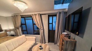 A modern living room with a beige sofa, a wooden coffee table, a wooden sideboard, and a large window with curtains. - Bryanston Palms Accommodation  (Sandton)