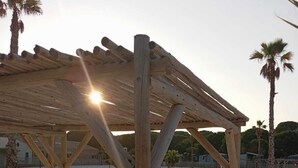 Un abri de plage en bois, doté d’un toit incliné soutenu par des poutres en bois, se dresse sur une plage de sable, avec des palmiers en arrière-plan.