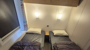 Une chambre de taille modeste, équipée de deux lits, chacun avec un oreiller et une couverture. Des lampes murales sont fixées au-dessus des lits, et une petite table se trouve entre les deux.