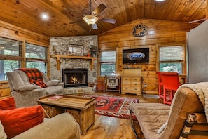 Un intérieur de chalet chaleureux avec une cheminée, des murs en bois, un mur d’accent en pierre et un téléviseur.