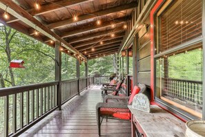 Une véranda couverte avec un plancher en bois, une balustrade, des meubles en osier et des guirlandes lumineuses.