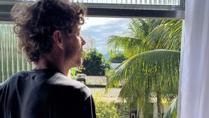 Une personne se tient près d’une fenêtre, d’où l’on aperçoit un jardin avec une piscine et des palmiers.