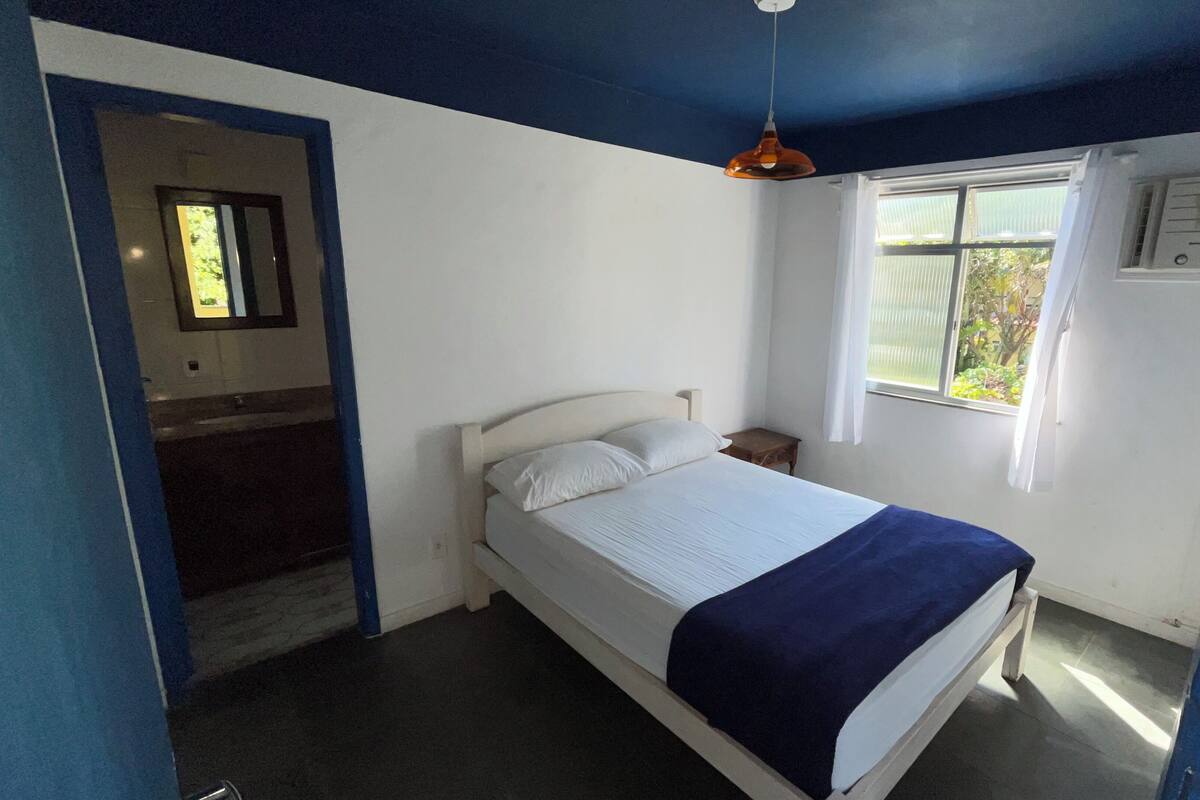 A bedroom with a bed, a bedside table, a hanging light fixture, a window with curtains, and a door with an air conditioning unit.