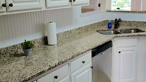 A kitchen with white cabinets, granite countertops, a dishwasher, and a window with a view of greenery.