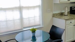 A small dining area with a round glass table, two black chairs, a potted plant, and white cabinets in the background.