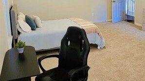 A bedroom with a bed, a desk with a chair, a ceiling fan, and a TV on the wall.
