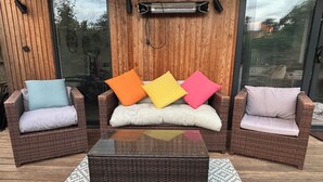 A wicker sofa with colorful cushions, a dark wicker coffee table, and a glass wall with a wooden panel backdrop.