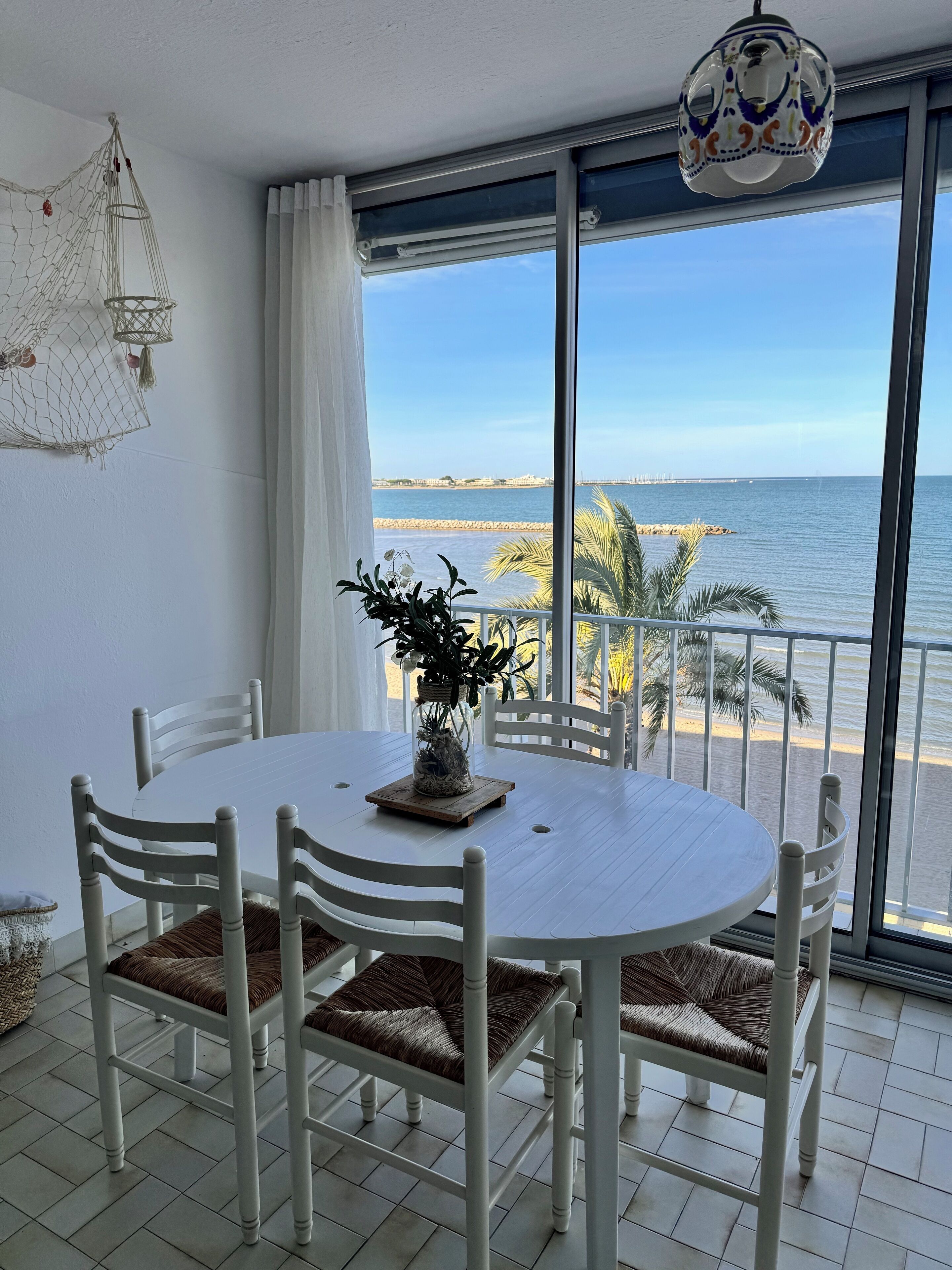 A dining area with a white table and chairs, a hanging light fixture, and a view of the beach through large windows.